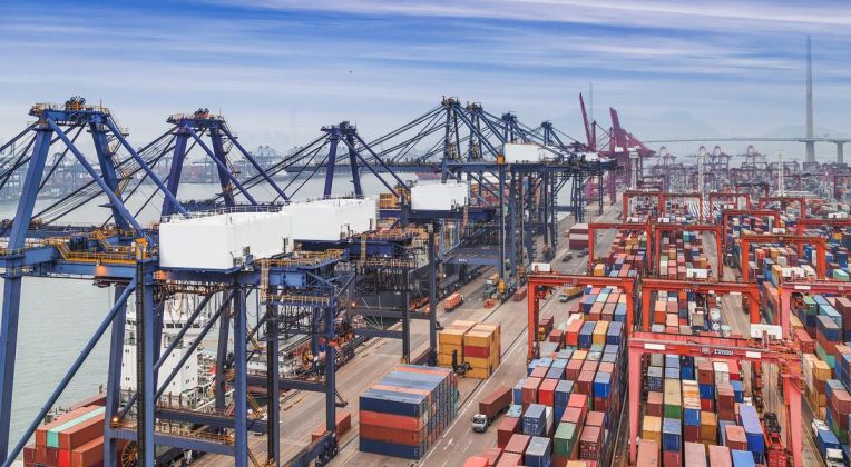 Panorama view of a container terminal with rows of containers, modern port cranes, and a coastline in the background.