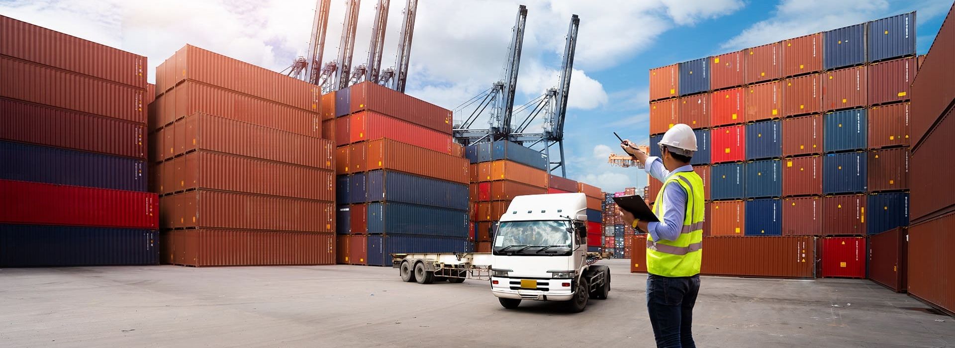 A port worker in a safety vest and helmet with a clipboard coordinating operations at a container terminal, surrounded by stacked containers, a truck, and an airplane in the sky.