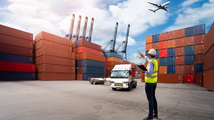 A port worker in a safety vest and helmet with a clipboard coordinating operations at a container terminal, surrounded by stacked containers, a truck, and an airplane in the sky.