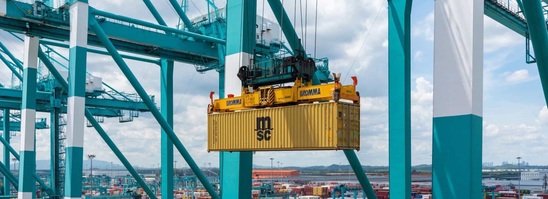 A yellow container being lifted by a crane in a container terminal, surrounded by stacked containers and modern port infrastructure.