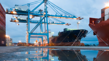 Rain clouds in the sky. Container terminal. Blue cranes. Container ship reflected in large puddle. Container stack on the left.