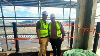 On-Site Partners in Terminal Transformation Two men (the one on the left is our colleague Holger) wearing safety vests and helmets standing on a construction site overlooking a container terminal in the background.