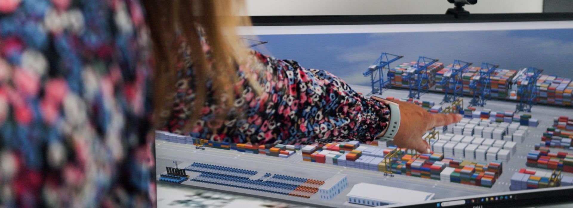A woman pointing at a screen displaying a 3D visualization of a container terminal represented as a digital twin.