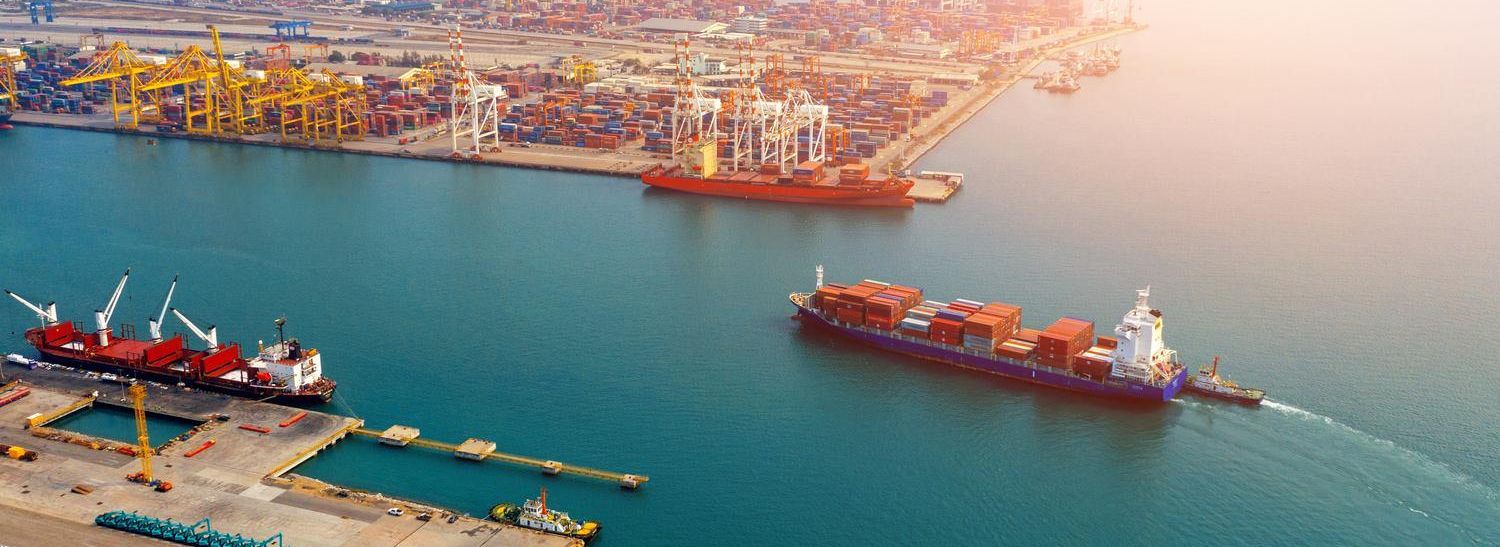 Aerial view of a container port with large cranes, cargo ships, and stacked containers in calm water, illuminated by the evening sun.