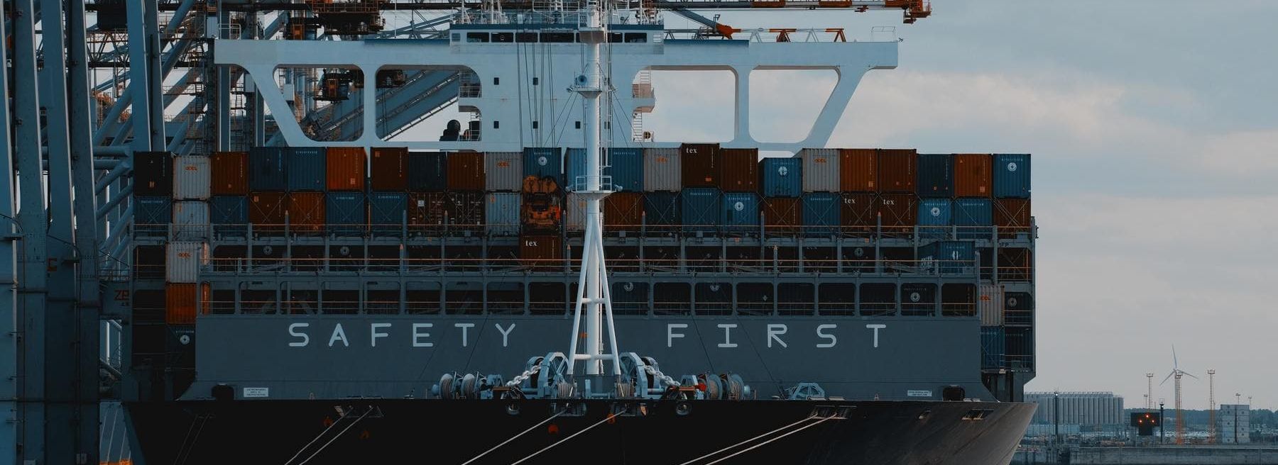 Close-up of a cargo ship with the text 'Safety First', surrounded by port cranes at a container terminal.