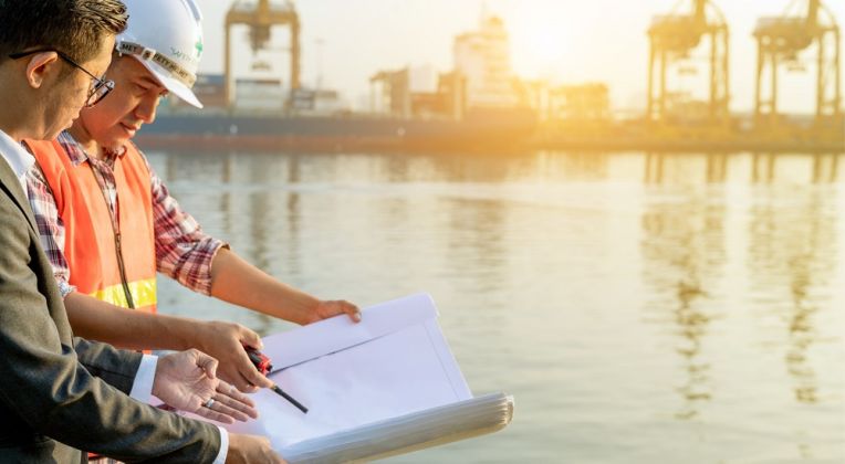 Two professionals, including an engineer in a safety vest and helmet, analyzing construction plans against the backdrop of a port with cranes and water in the golden morning sunlight.