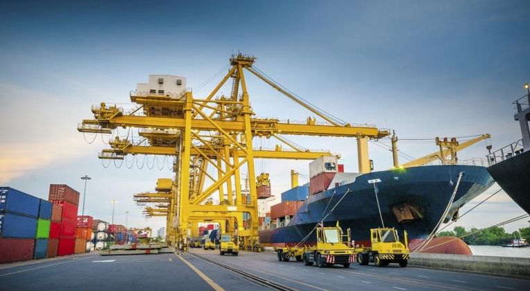 A cargo ship in a container terminal being unloaded by large yellow cranes while transport vehicles move containers.