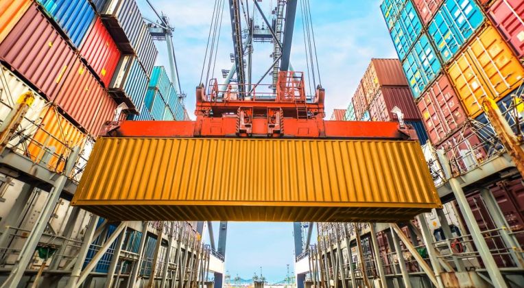 Close-up of a yellow container being moved by a crane in a container terminal, surrounded by stacked containers.