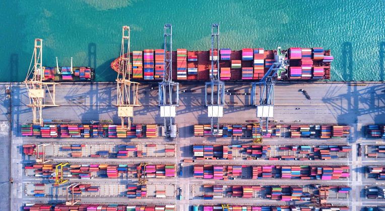 Aerial view of a container port with stacked containers, large cranes, and a cargo ship in turquoise water.