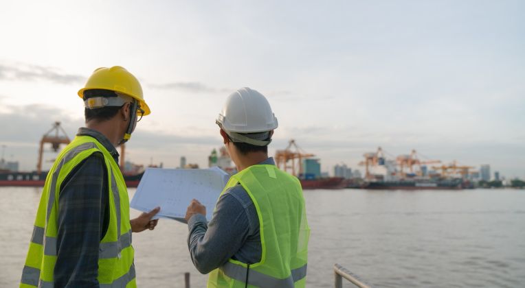 Two engineers wearing safety vests and helmets stand at a harbor reviewing construction plans, with cranes and container ships in the background.