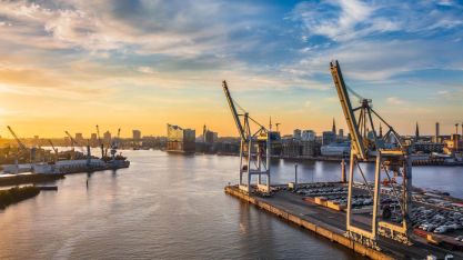 View of the Port of Hamburg at sunset featuring container cranes along the Elbe River and the Elbphilharmonie in the background.