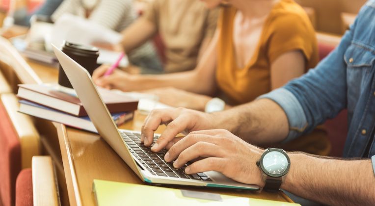 Close-up of a student working on a laptop while other students take notes in the background.
